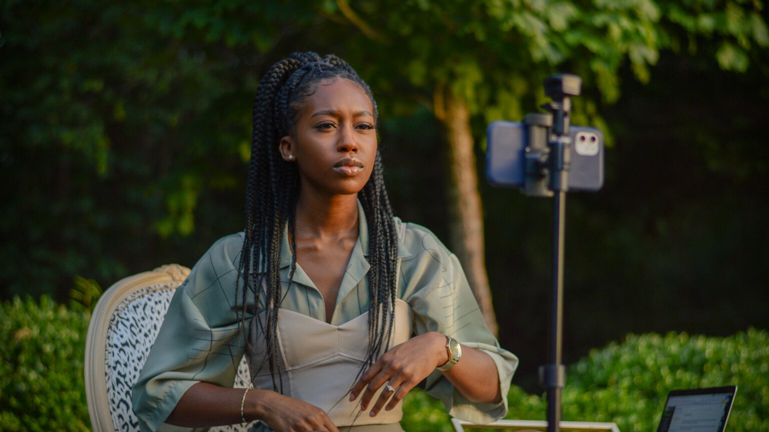 Lynae sits on a chair outside, surrounded by greenery. She is looking at a phone set up on a tripod. 