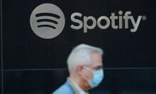 A Spotify logo is visible in the center of the image while a man wearing a surgical mask is visible and out of focus in the foreground.