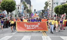 A group of people march down a street during a Pride parade. They are holding a large orange Trevor Project banner. 