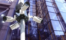 Several street surveillance cameras in front of a glass building.