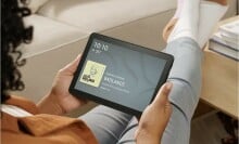 a person sits with legs extended on a coffee table while holding the fire HD 8 tablet in their hands. The display screen of the tablet shows the time, temperature, and the current song playing