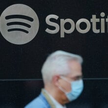 A Spotify logo is visible in the center of the image while a man wearing a surgical mask is visible and out of focus in the foreground.