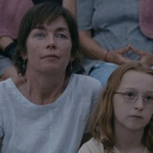 A woman in a white t-shirt and a young girl in glasses sit together on the grass.