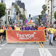 A group of people march down a street during a Pride parade. They are holding a large orange Trevor Project banner. 