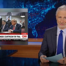 A man in a suit sits behind a talk show desk. In the top-left is an image from Trump's trial.