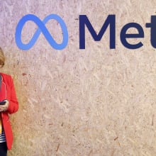 A woman in a red blazer leans against a wall with the Meta logo on it and looks down at her phone.