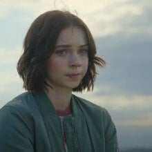 A teenage girl stands in front of a blue sky backdrop, looking worried.