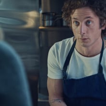 Carmy from "The Bear" in a restaurant kitchen, wearing a blue apron and white t-shirt.