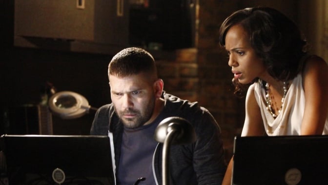 A man sits behind a desk, staring at two computer screens, while a woman stands behind him.