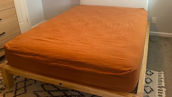 Mattress with burnt orange sheets atop a wood platform bed frame with white pillow board