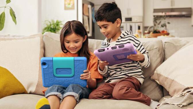 Two kids sitting on the couch and playing on their Amazon Fire 7 Kids tablets with smiles on their faces.