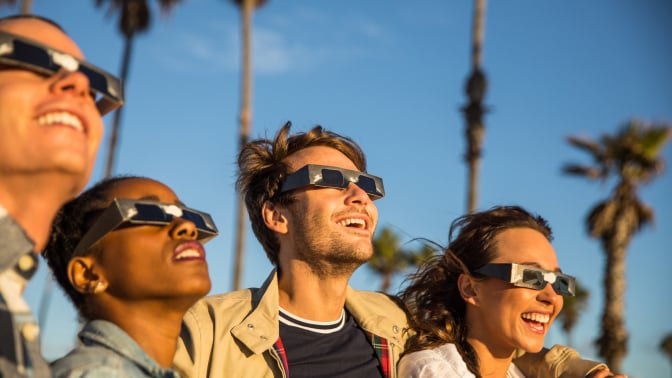 four people wearing eclipse glasses smile while looking into the sky