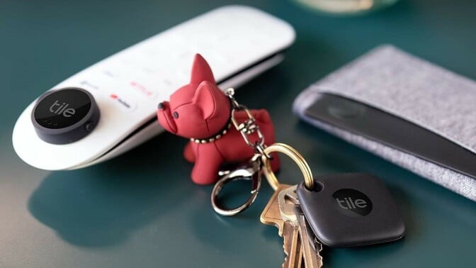 a side table includes a remote control, keys with a red dog keychain, and a wallet. All items have a tile bluetooth tracker attached.