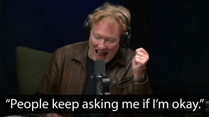 A man sits in a dark room at a table in front of a microphone.