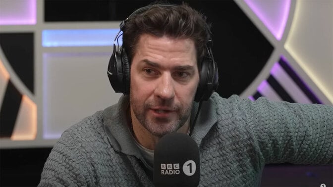 A man sits in a radio booth in front of a microphone, wearing a headset.