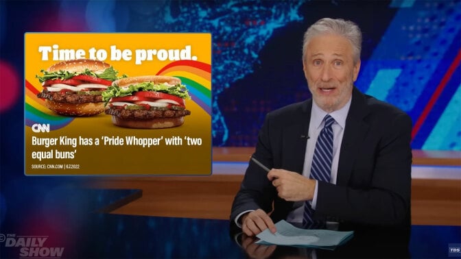 A man sits behind a talk show desk. In the top-left is a Burger King advertisement for Pride.