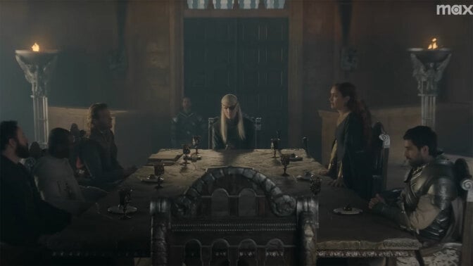 A group of people in medieval clothes sit around a large table in a gloomy room.