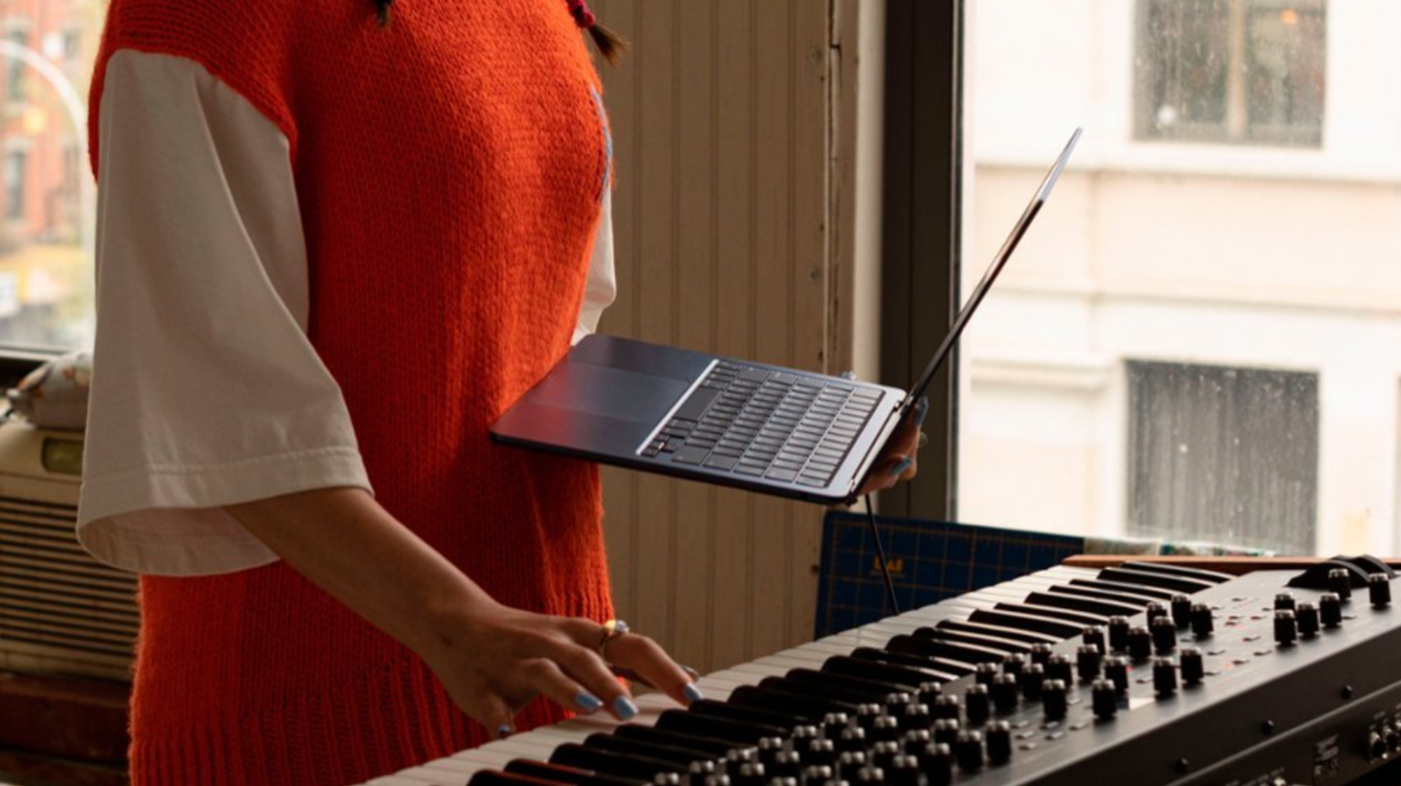 a woman holding an m2 apple macbook air in one hand while playing a keyboard with the other