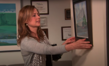 A woman (Jenna Fischer as Pam Beesly) removing a framed watercolor painting of an office building from the wall in a scene of "The Office."