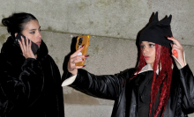 Two young women on a sidewalk, one talking on her cell phone, the other taking a selfie while wearing a black hat with cat ears