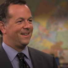 A man (David Costabile as the banker on "The Office") standing in an office wearing a suit and smiling.