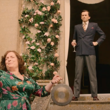 A woman bashes a gong while a man in a suit stands in a doorway with a flute of bubbly.