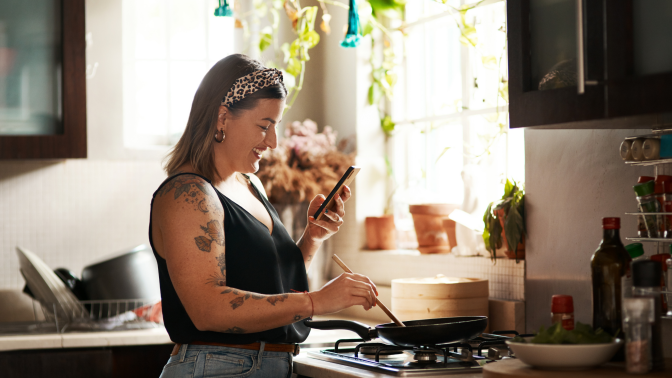 Happy, woman cooking food with smartphone and in kitchen of a home. Dinner or lunch, multitasking and plus size, natural person with tattoo on cellphone following recipe motivation for healthy diet